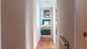 Un pasillo estrecho con suelo de madera en dibujo de espiga que conduce a un dormitorio con cabecera color turquesa y ropa de cama blanca.