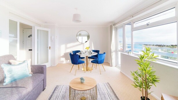 A modern living room with a grey sofa, a round coffee table, and a dining table with blue chairs. There is a large window with a view of the sea.