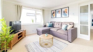 A modern living room with a sectional sofa, a television on a wooden stand, a round coffee table, and a large window with curtains.