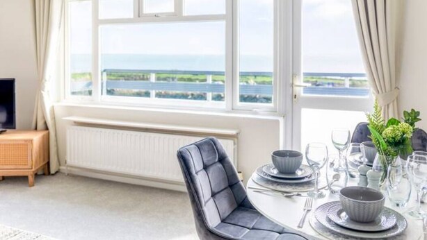 A dining area with a table set for four, a view of the sea through large windows, and a modern chair. - Large Sea View 2 bedroom Sleeps 6 - Free Parking (Bournemouth)
