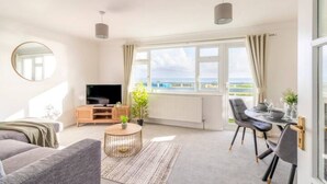A modern living room with a large window, a flat-screen TV, a sofa, a coffee table, and a dining area with chairs and a table. - Large Sea View 2 bedroom Sleeps 6 - Free Parking (Bournemouth)