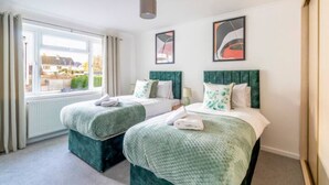 A hotel room with two beds, each with green headboards and matching green bedding. There is a window with curtains, a radiator below the window, and two abstract paintings on the wall. - Large Sea View 2 bedroom Sleeps 6 - Free Parking (Bournemouth)