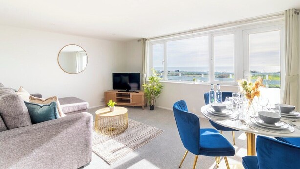 Un salon moderne avec un canapé, un téléviseur, une table à manger pour quatre personnes et une grande fenêtre donnant sur la mer.