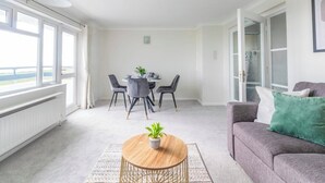Een moderne woonkamer met een grijze bank, een ronde salontafel met een plant, een eethoek met een tafel en stoelen, en grote ramen met uitzicht.