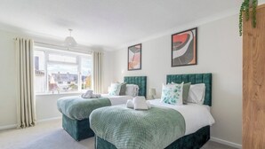 Hotelkamer met twee bedden, elk met groene hoofdborden en passend beddengoed. Groot raam met gordijnen, hangplant en abstracte kunst aan de wanden.