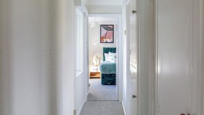 Een smalle gang die leidt naar een slaapkamer met een bed, een nachtkastje en een lamp.
