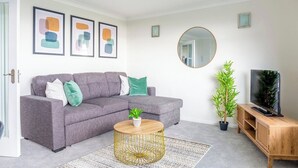 Een woonkamer met een grijze bank, een ronde salontafel, een tv op een houten standaard en twee planten in potten.