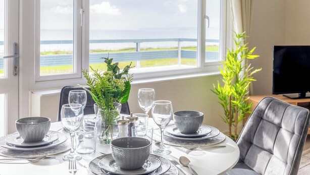 A dining table set for six with a view of the sea through large windows. - Panoramic Sea View Apartment Sleeps 4-Free Parking (Bournemouth)
