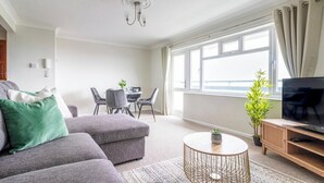 A modern living room with a grey sofa, a round coffee table, a TV on a wooden stand, and a dining area with a table and chairs. - Panoramic Sea View Apartment Sleeps 4-Free Parking (Bournemouth)