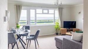 A living room with a large window, a television, and a dining area with chairs and a table. - Panoramic Sea View Apartment Sleeps 4-Free Parking (Bournemouth)