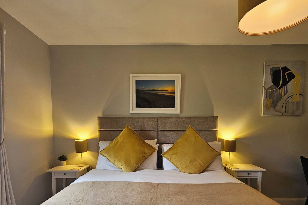 Habitación de hotel ordenada con una cama grande, dos mesitas de noche con lámparas, un cuadro en la pared y una pequeña mesa con una planta.