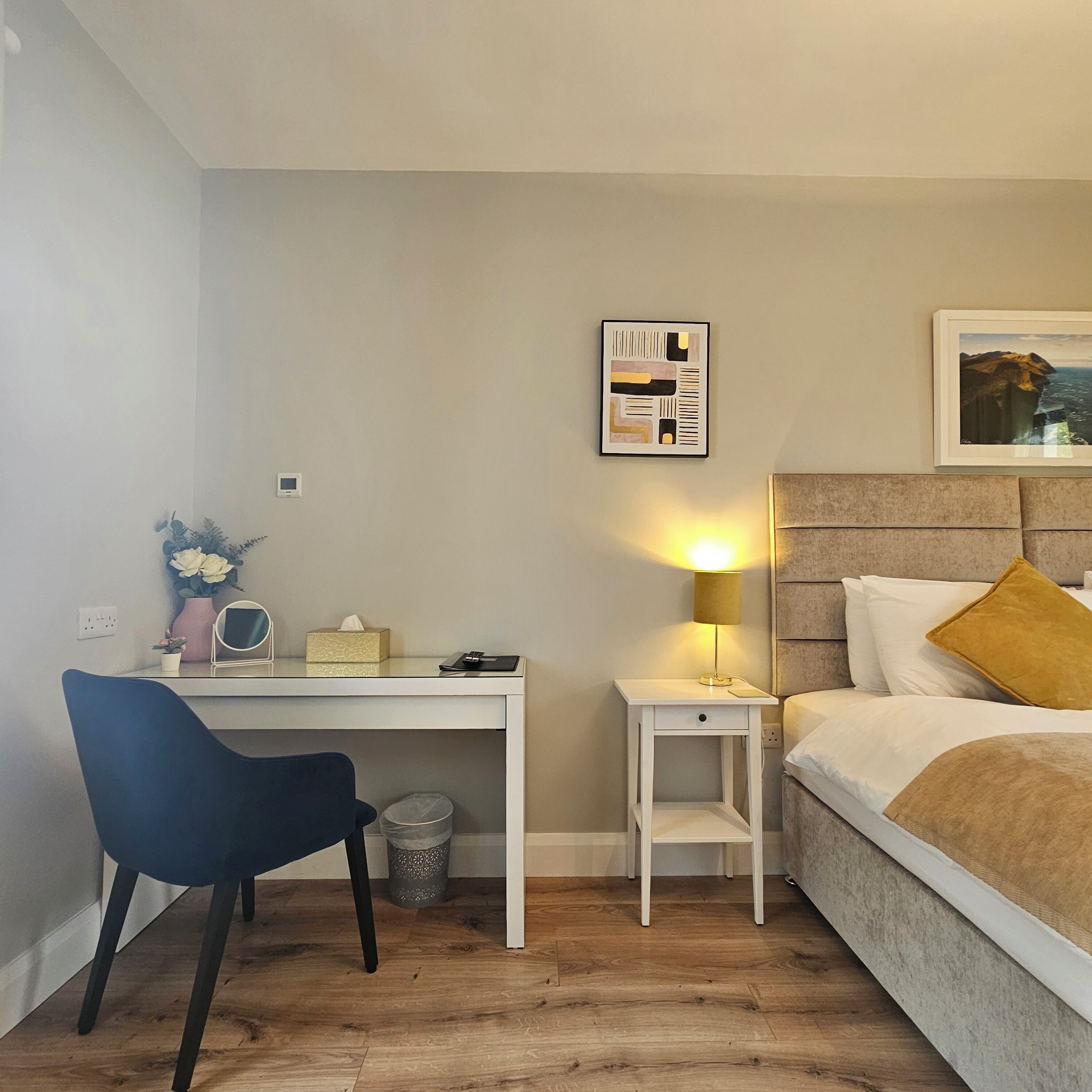 A hotel room with a desk, chair, bedside table with a lamp, a bed with pillows, and a framed picture on the wall.