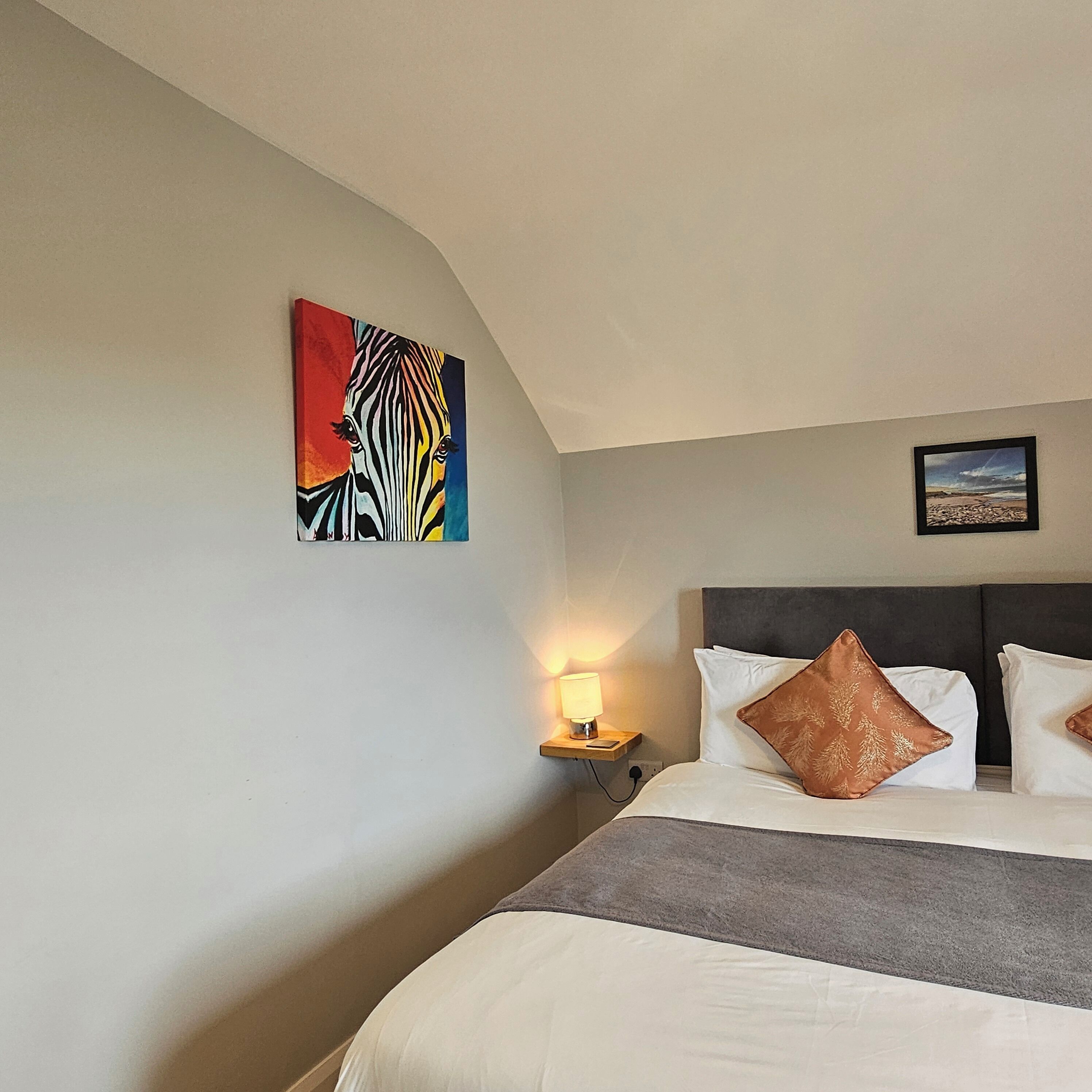 A hotel room with a bed, pillows, a nightstand with a lamp, and two wall art pieces.