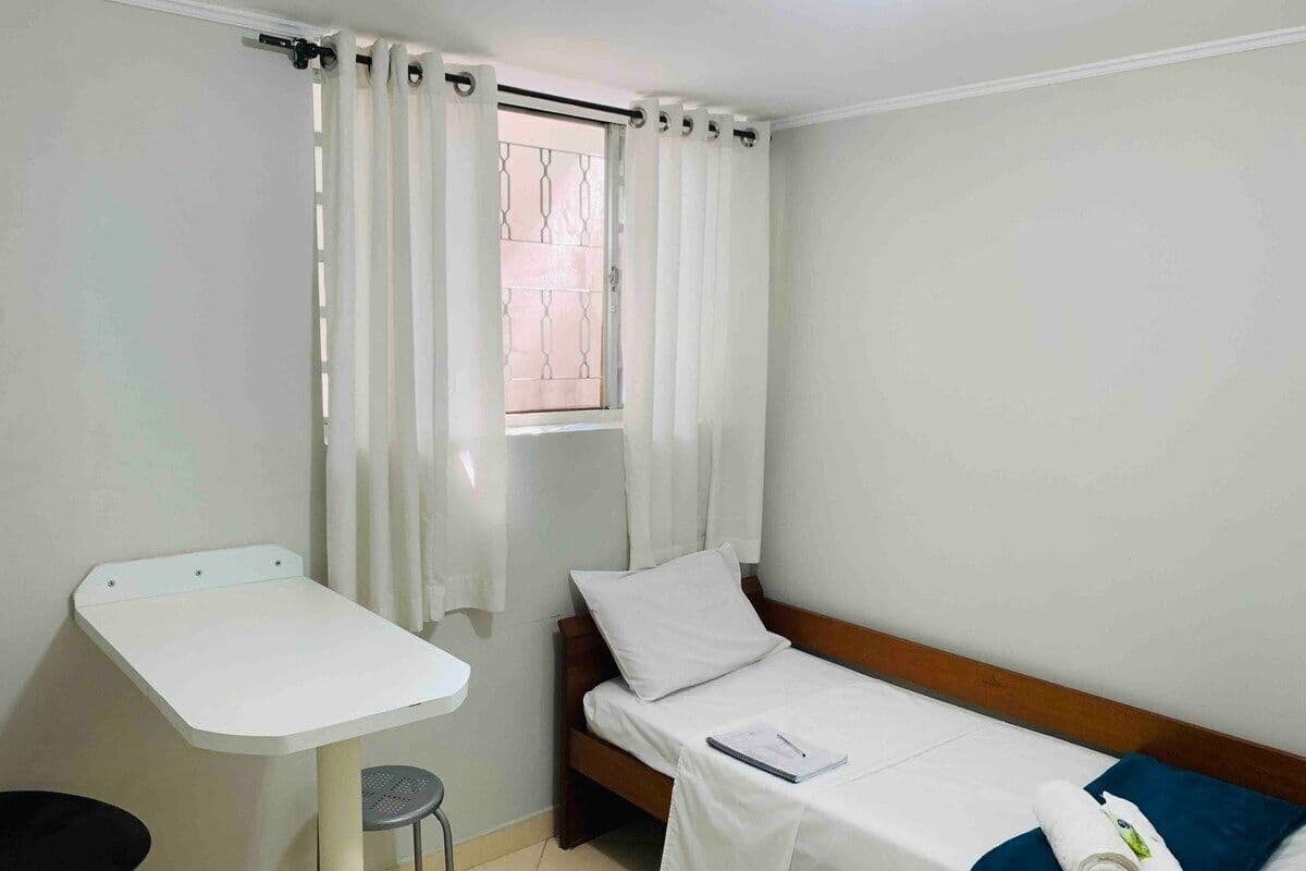 A small, clean room with a single bed, a small table, and a window with curtains.