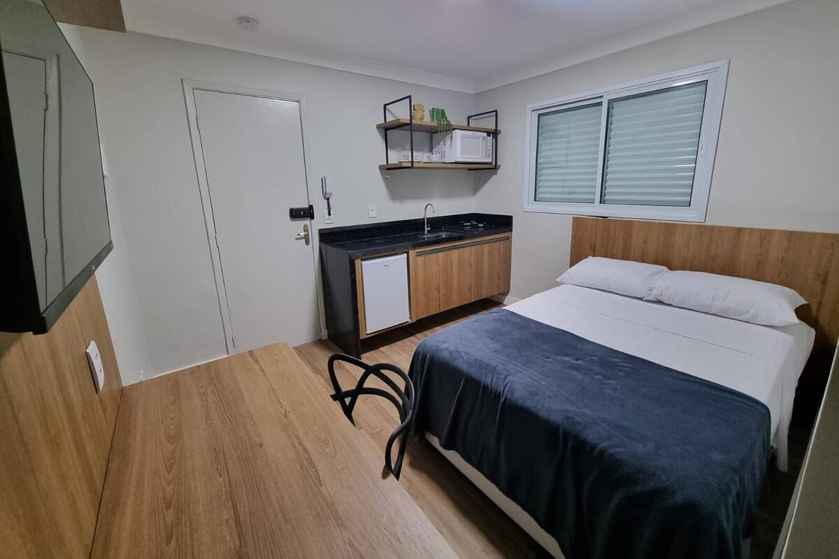 A compact bedroom with a bed, a small kitchenette, and a window with blinds.
