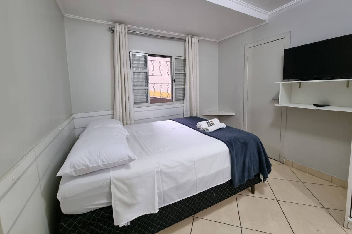A bedroom with a bed, a window with curtains, a television on a shelf, and a tiled floor.