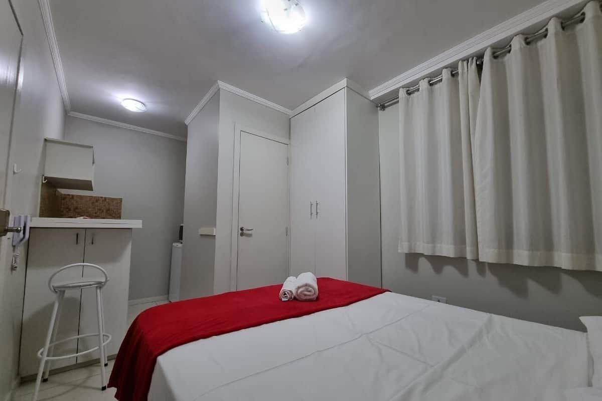 A hotel room with a bed, a red blanket, white curtains, a white wardrobe, and a small stool.