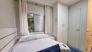 Un dormitorio con cama, cortinas blancas, una ventana con persianas, un armario y una toalla sobre la cama.