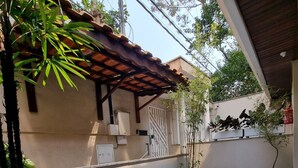 Un patio techado con piso de baldosas, un banco de madera, plantas en macetas y un portón blanco.