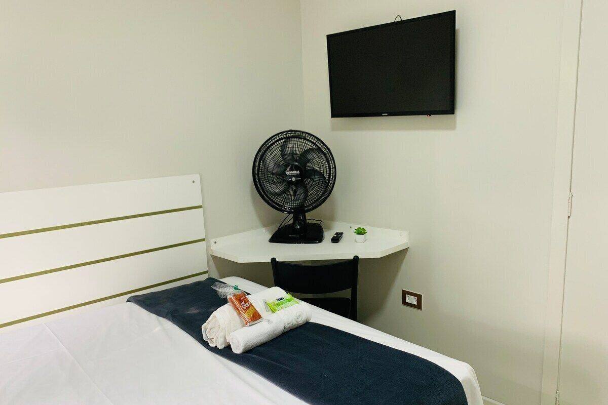 A hotel room with a bed, a desk, a chair, a fan, and a TV on the wall.