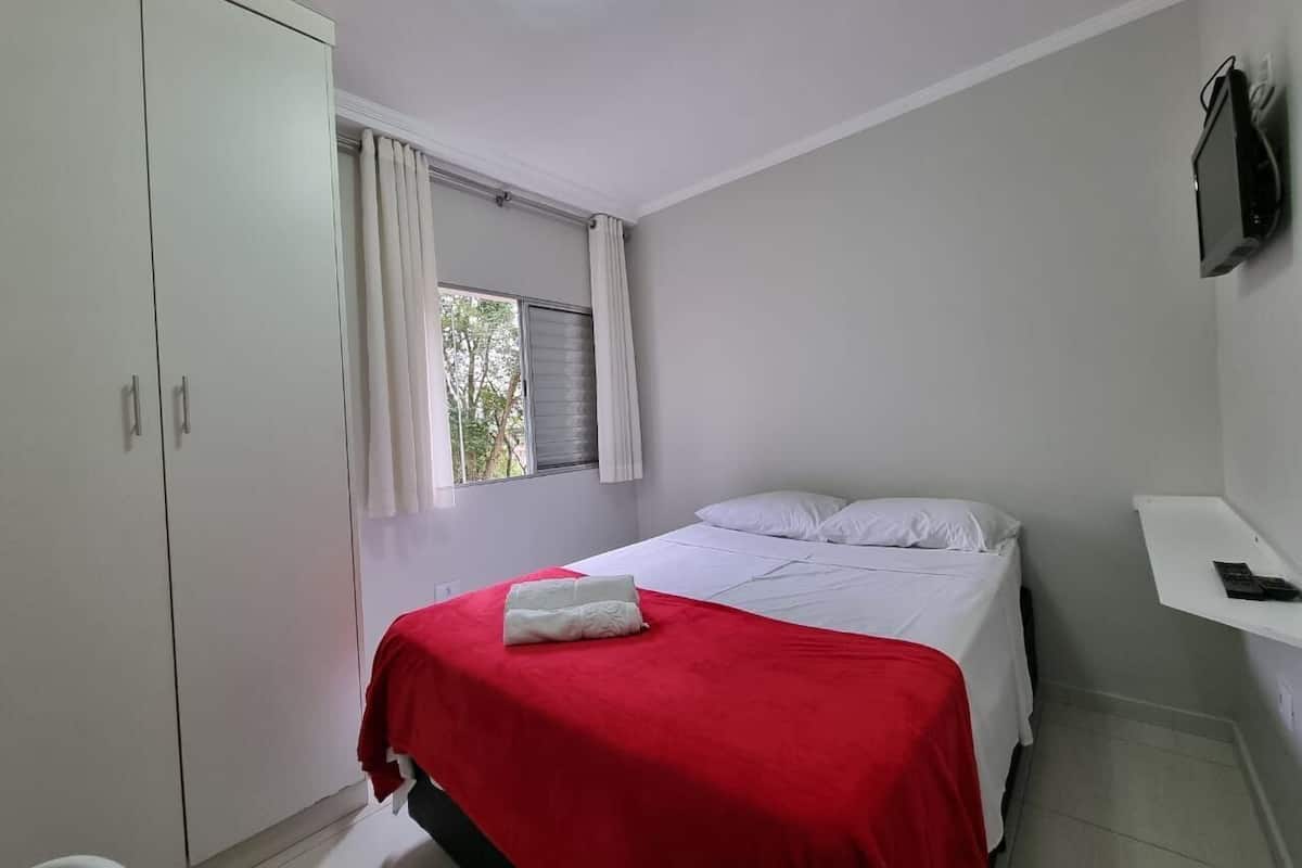 A bedroom with a bed, white curtains, a window, a wardrobe, and a flat-screen TV.
