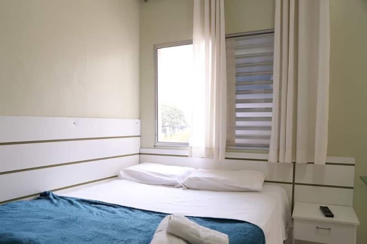 A simple, clean hotel room with a single bed, a nightstand with a phone, and a window with blinds.