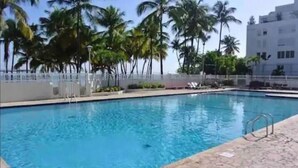 A swimming pool with a tiled edge, surrounded by palm trees and a white building in the background. - Sleep with the sound of waves! (Carolina)
