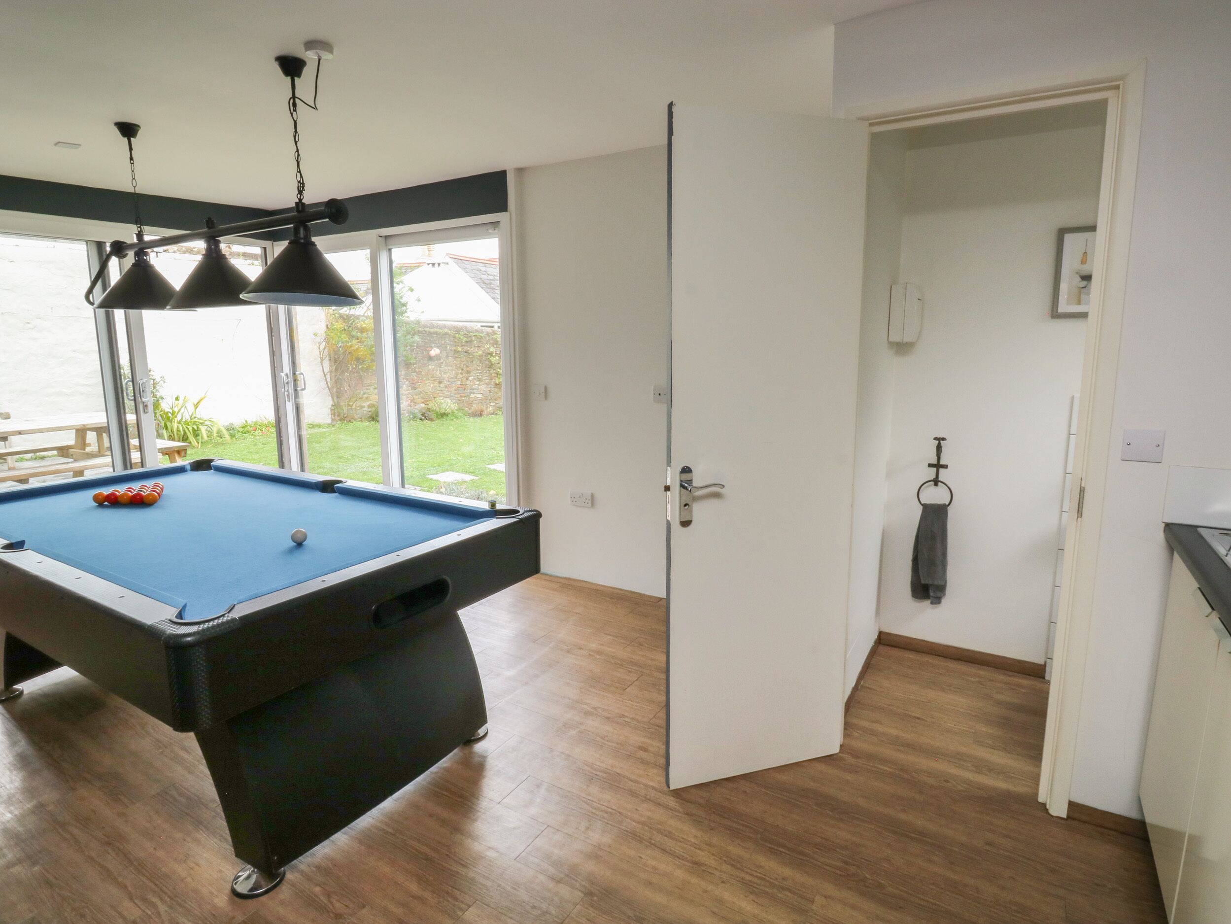 A pool table with three hanging lamps, a door leading to a balcony, and a view of a garden.