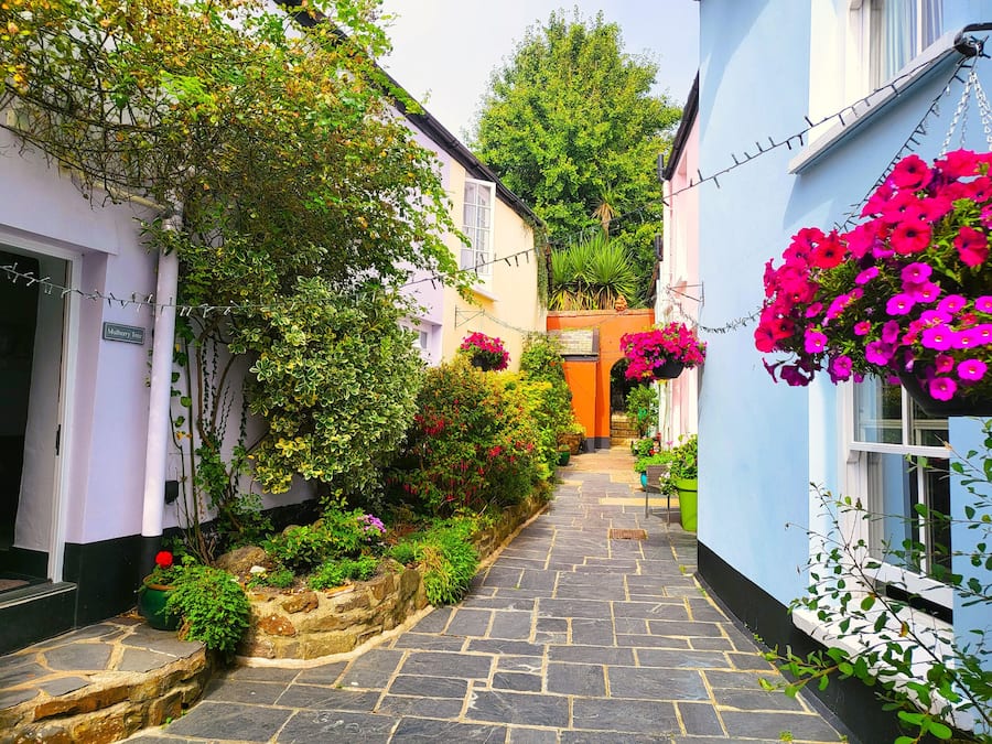 A narrow stone-paved alleyway flanked by buildings with vibrant flower boxes and lush greenery.
