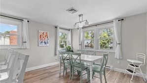 Een eetgedeelte met een tafel en stoelen, grote ramen voorzien van gordijnen, en een ventilatieopening in het plafond.