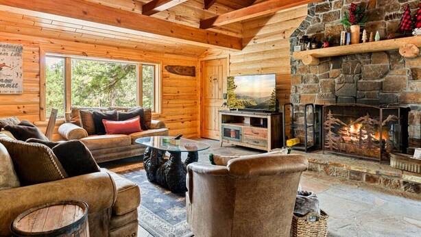 Interior acogedor de una cabaña de troncos con chimenea de piedra, mobiliario de madera y un ventanal con vistas a la vegetación.