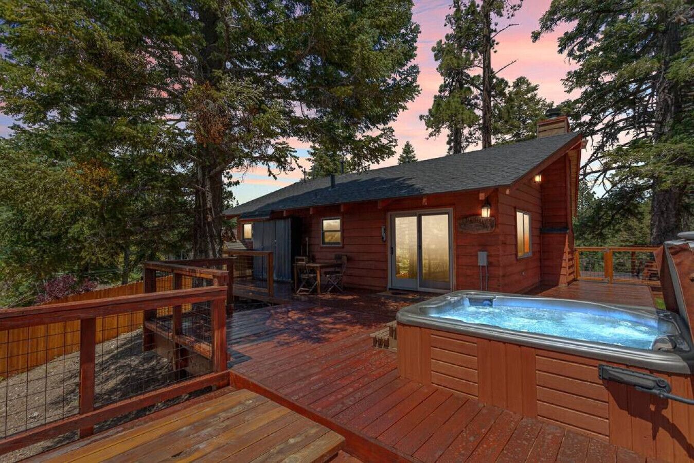 A wooden cabin with a hot tub, deck, and a covered patio area surrounded by trees.