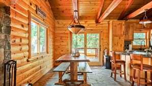 Interior de una cabaña de troncos con suelo de piedra, paredes de madera, chimenea de piedra, mesa de comedor de madera con bancos y una zona de cocina con fregadero y taburetes de bar.