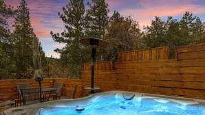 A hot tub surrounded by wooden deck furniture, a table, and chairs, with a backdrop of trees and a sunset sky. - Silver Linings+Hot Tub+Pet Friendly+EV+Moonridge (Big Bear Lake)