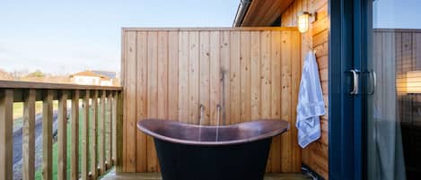Una terraza de madera con una bañera independiente, una toalla colgada en la pared y un vidrio que da a un área cubierta.