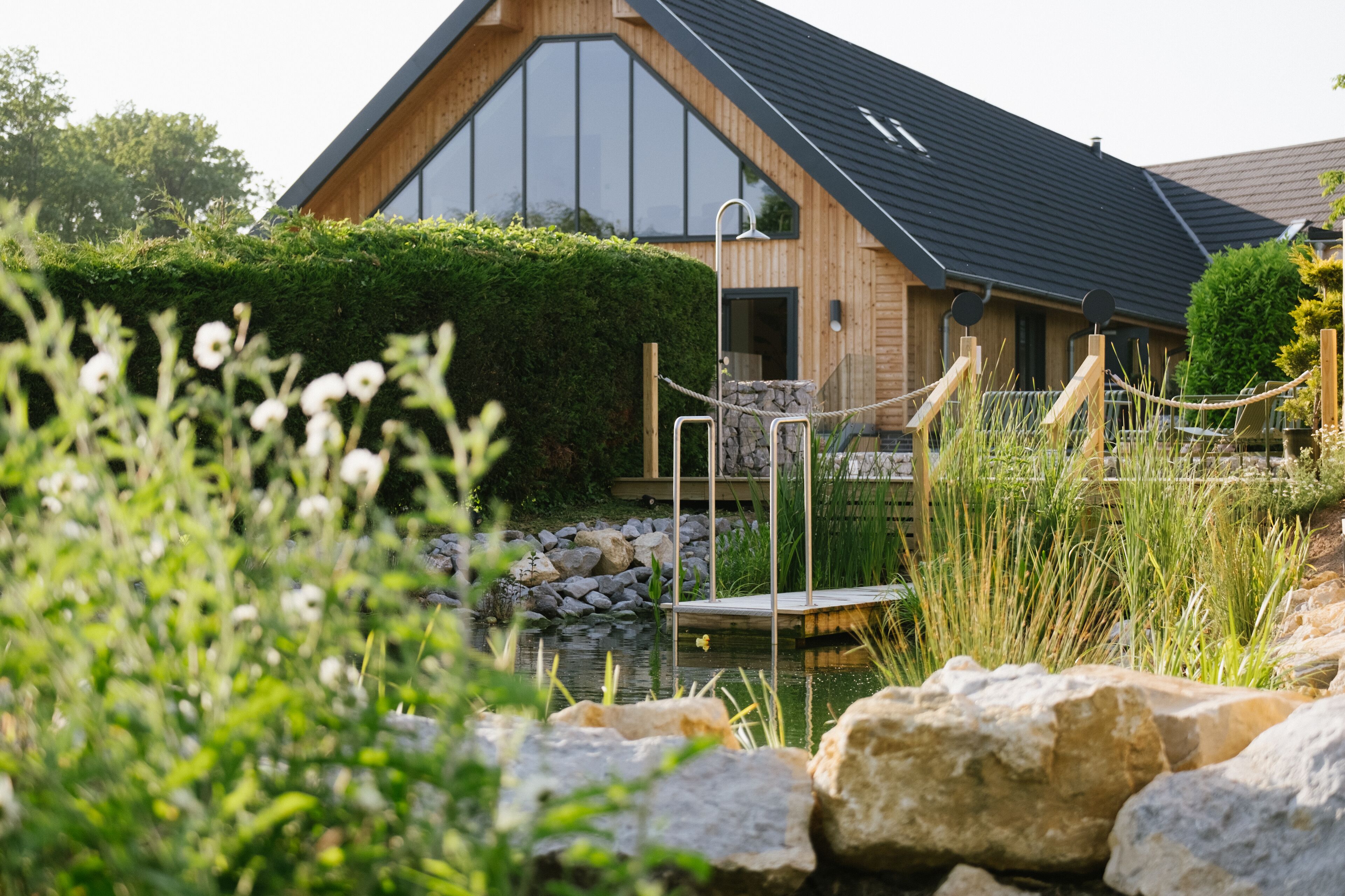Une maison moderne en bois, dotée d’une grande fenêtre, d’une terrasse en bois et d’un étang bordé de rochers.