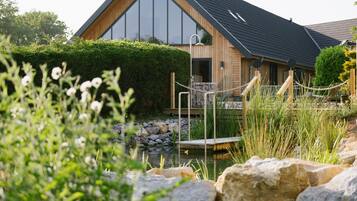 A modern wooden house with a large window, a wooden deck, and a pond with rocks.