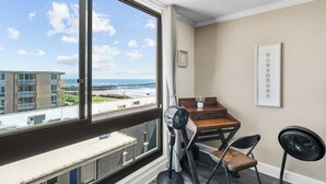 Une pièce avec une grande fenêtre, un bureau, deux chaises, un ventilateur et un tableau encadré au mur.