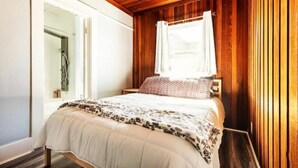Un dormitorio con techo de madera, una cama con manta estampada y una ventana con cortina blanca.