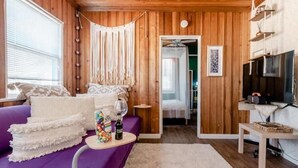 A cozy room with wooden walls, a purple sofa, a small table with a bottle and snacks, and a TV on a shelf. - Tuluminati:BohoChic Condo Steps from Ocean and Bay (San Diego)