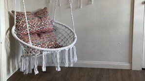 A macramé hanging chair with colorful cushions in a room with a wooden floor and a plain wall. - Tuluminati:BohoChic Condo Steps from Ocean and Bay (San Diego)