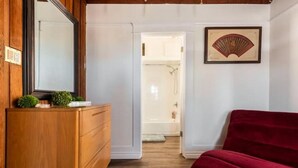 A bedroom with a wooden dresser, a red chair, a framed artwork on the wall, and a bathroom visible through an open door. - Luxe Thai-Inspired Condo Steps to Beach w/ Parking (San Diego)