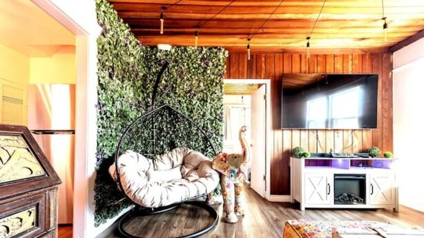 A cozy living room with a wooden ceiling, a large flat-screen TV, a plush chair, a small table, and a decorative wall with greenery. - Luxe Thai-Inspired Condo Steps to Beach w/ Parking (San Diego)