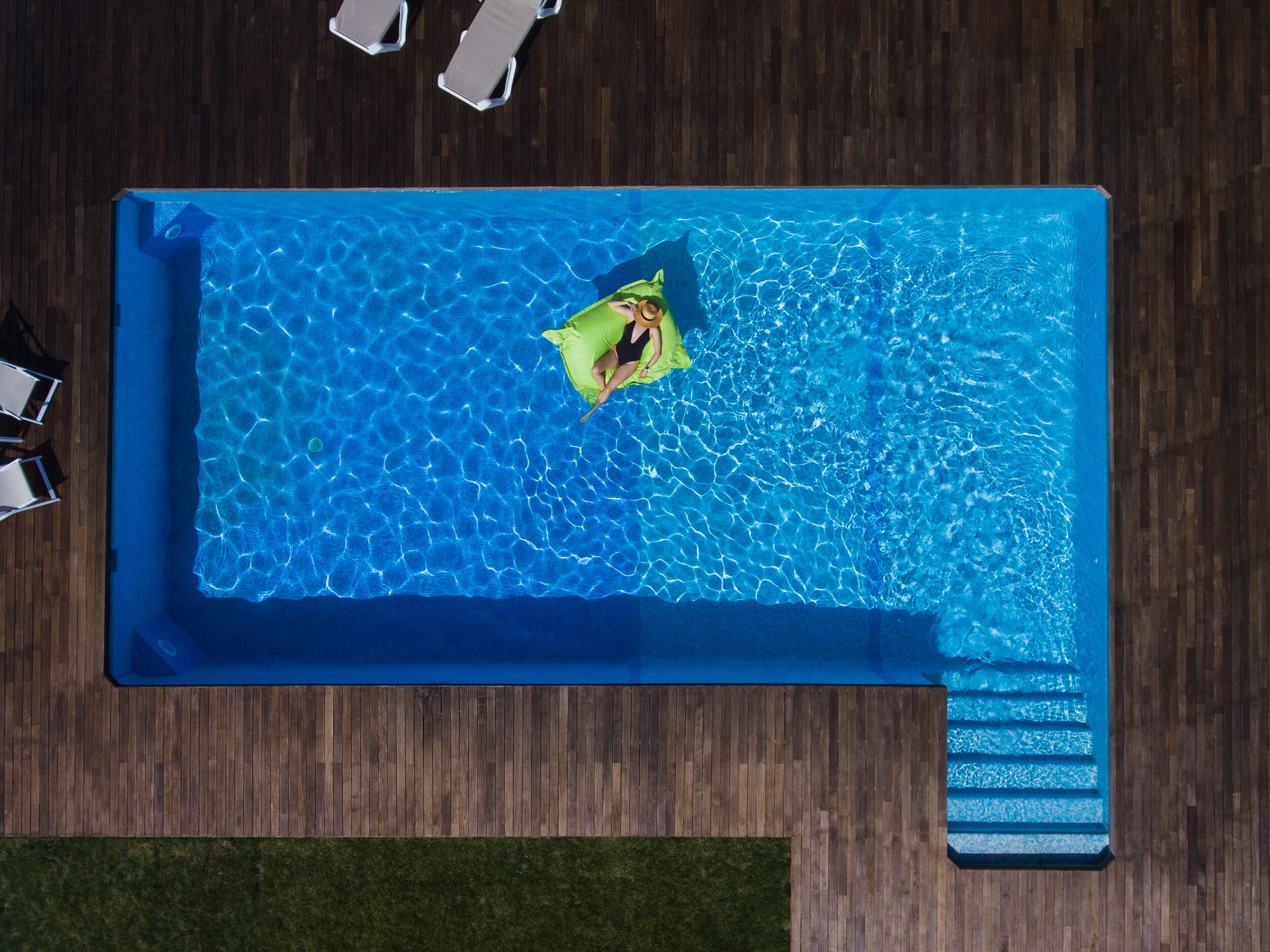 Una piscina rectangular con agua azul cristalina, un deck de madera alrededor y una zona verde con césped al fondo.