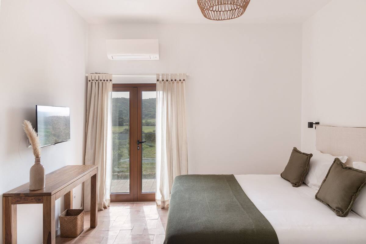 A bedroom with a bed, a bedside table, a TV, and a window with curtains.