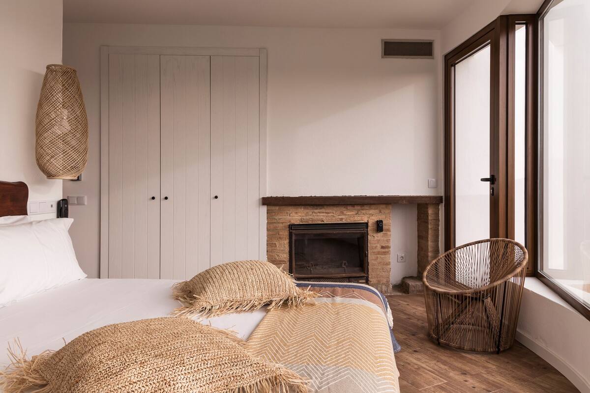 A bedroom with a bed, a woven chair, a fireplace, and a large window.