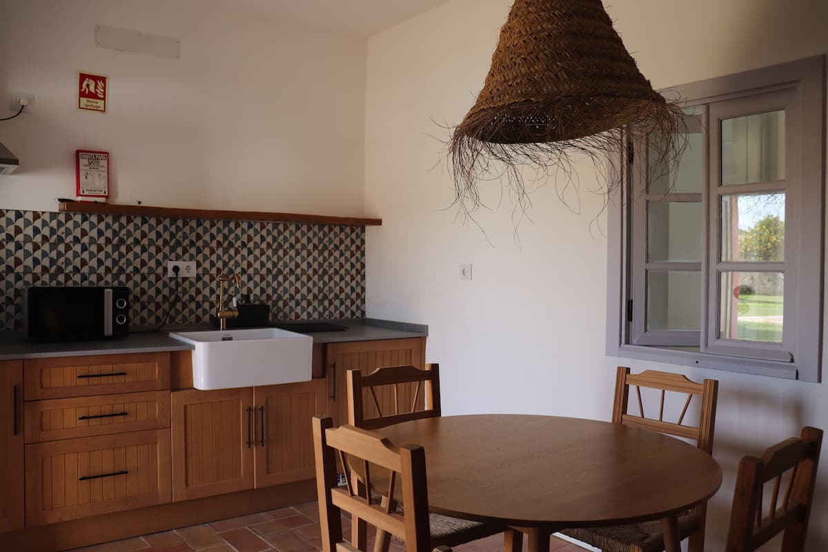 A kitchen with wooden cabinets, a microwave, and a sink. There is a round wooden table with chairs. A woven hanging light fixture is above the table.