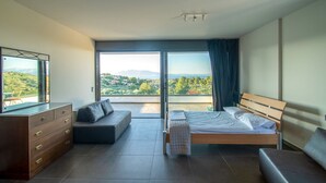 Ein modernes Schlafzimmer mit Bett, Sofa, Kommode und einem großen Fenster mit Blick ins Grüne.