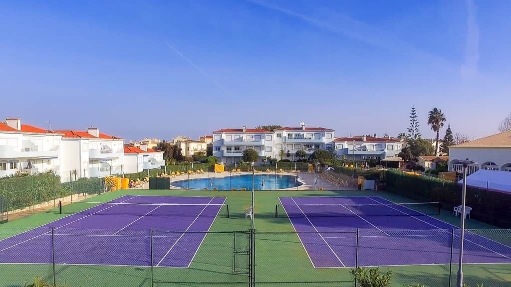 Een tennisbaan met een zwembad op de achtergrond, omringd door woongebouwen.
