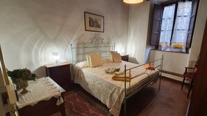 Ein Schlafzimmer mit Metallbettgestell, einem Nachttisch mit Lampe, einem Fenster mit Vorhängen und einem gerahmten Bild an der Wand.